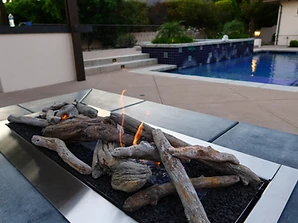 Linear driftwood gas fire pit on an outdoor patio with swimming pool in the background